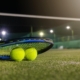 Image of a racket and balls laying on a tennis court being lit up at night.